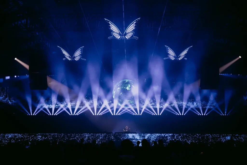 Palco indoor iluminado de azul, com borboletas de luz suspensas e um globo ao fundo